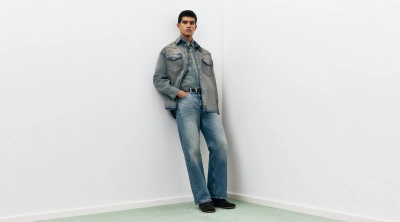 Model Bodhi Heeck leaning against a white studio wall wearing a washed denim jacket, denim shirt, and relaxed blue jeans.
