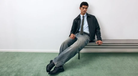Model Bodhi Heeck wearing a black leather jacket, striped shirt, tie, and faded wide-leg jeans seated on a bench.
