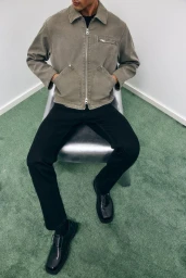 Model seated on a metallic chair in a green carpeted room wearing a taupe zip jacket and black trousers