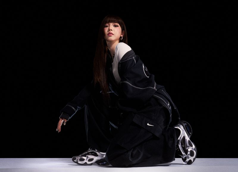 Model crouching in a black Nike tracksuit and metallic silver sneakers against a dark studio background.