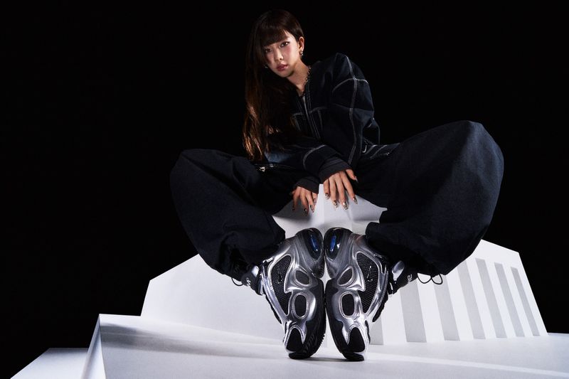 Portrait of an adult woman in a dark jacket and matching pants, seated on a white platform wearing metallic sneakers against black.