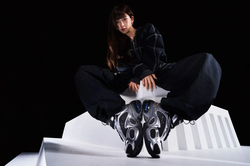 Portrait of an adult woman in a dark jacket and matching pants, seated on a white platform wearing metallic sneakers against black.