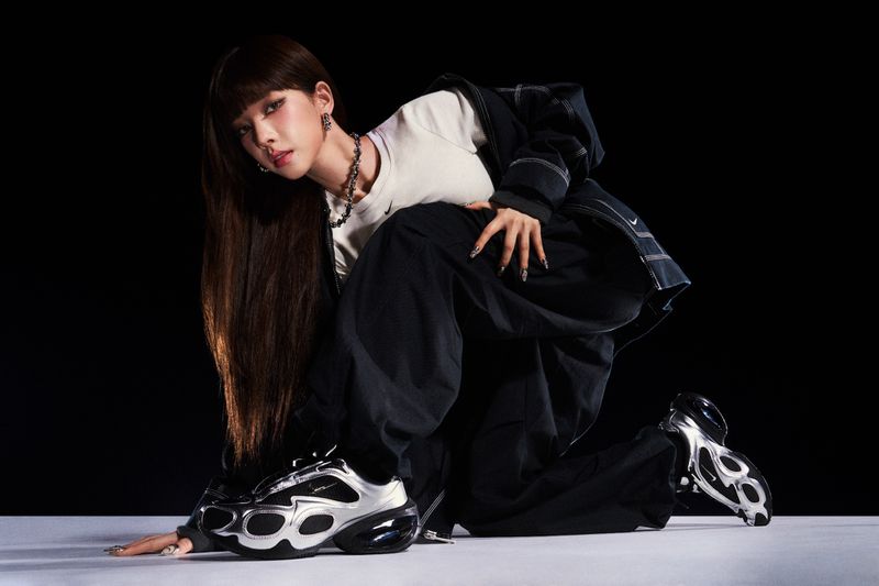 Woman posing in studio with silver and black sneakers, wearing a white shirt and dark pants and jacket against black backdrop.