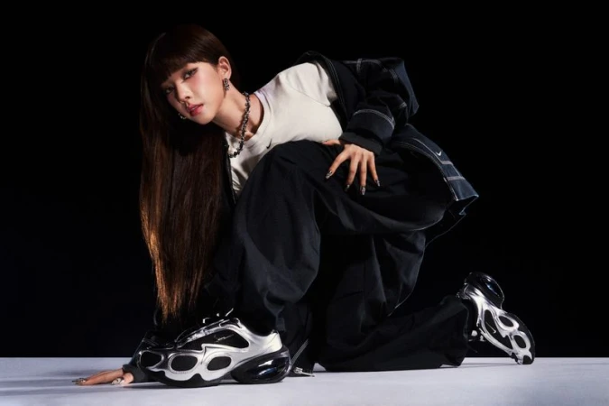 Woman posing in studio with silver and black sneakers, wearing a white shirt and dark pants and jacket against black backdrop.