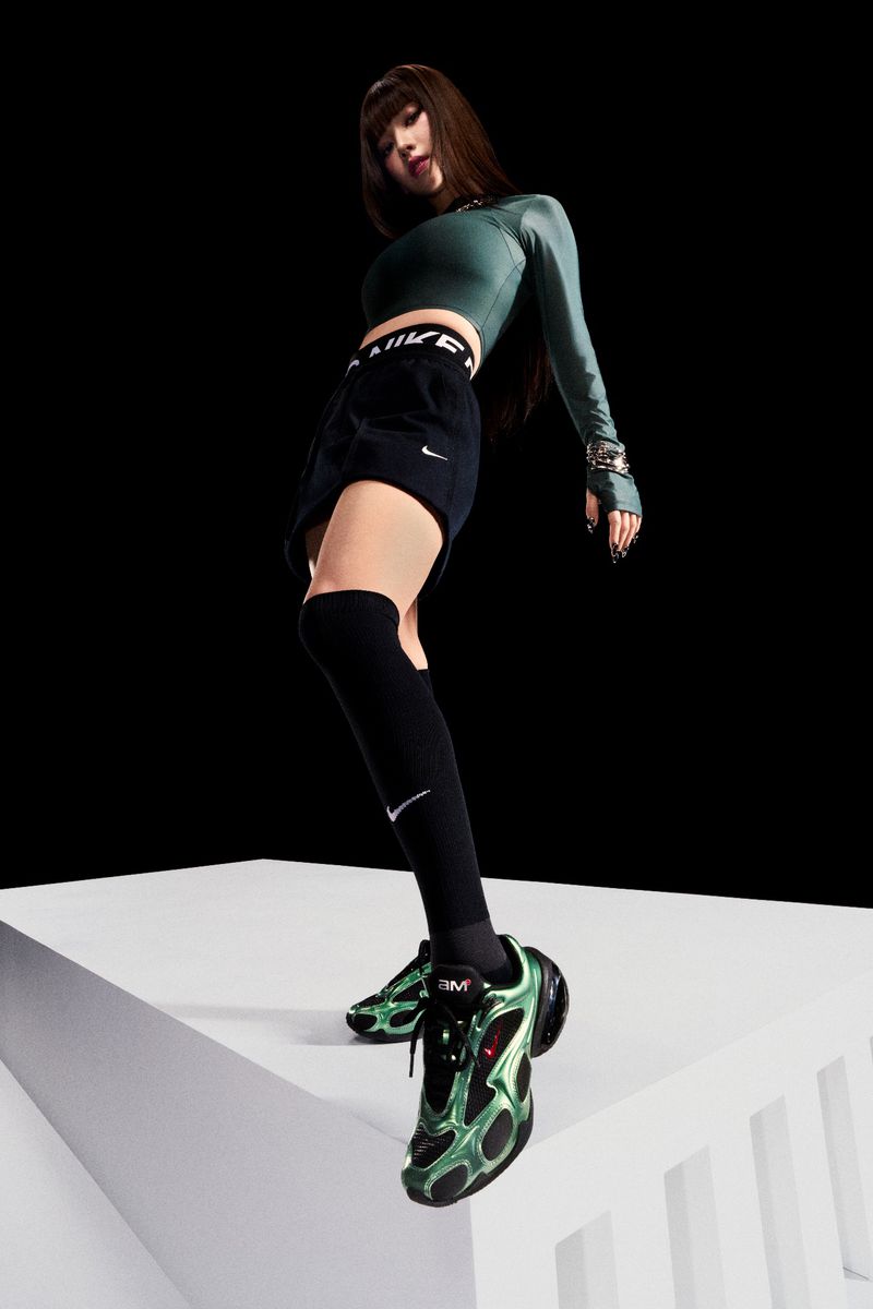 A woman stands on a white box in a photo studio, wearing a green long-sleeve crop top, black Nike shorts, black knee socks and green and black Nike sneakers.