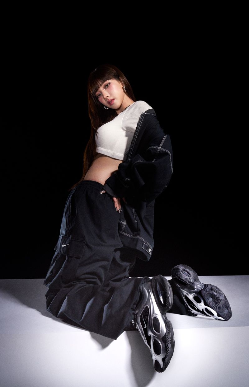 K-pop star Ningning poses kneeling in a white crop top, black cargo pants, and silver Nike sneakers against a dark studio background.