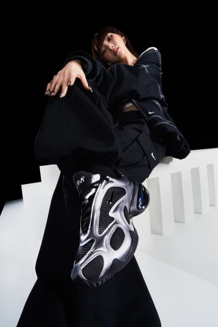 Stylish woman in black sportswear and silver sneakers poses on white steps against a black background in a studio shot.