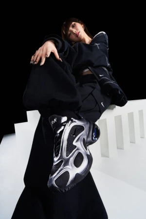 Stylish woman in black sportswear and silver sneakers poses on white steps against a black background in a studio shot.