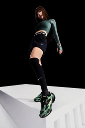 Low-angle studio shot of a woman wearing green crop top, black shorts, and metallic green Nike sneakers on a white block against a black backdrop