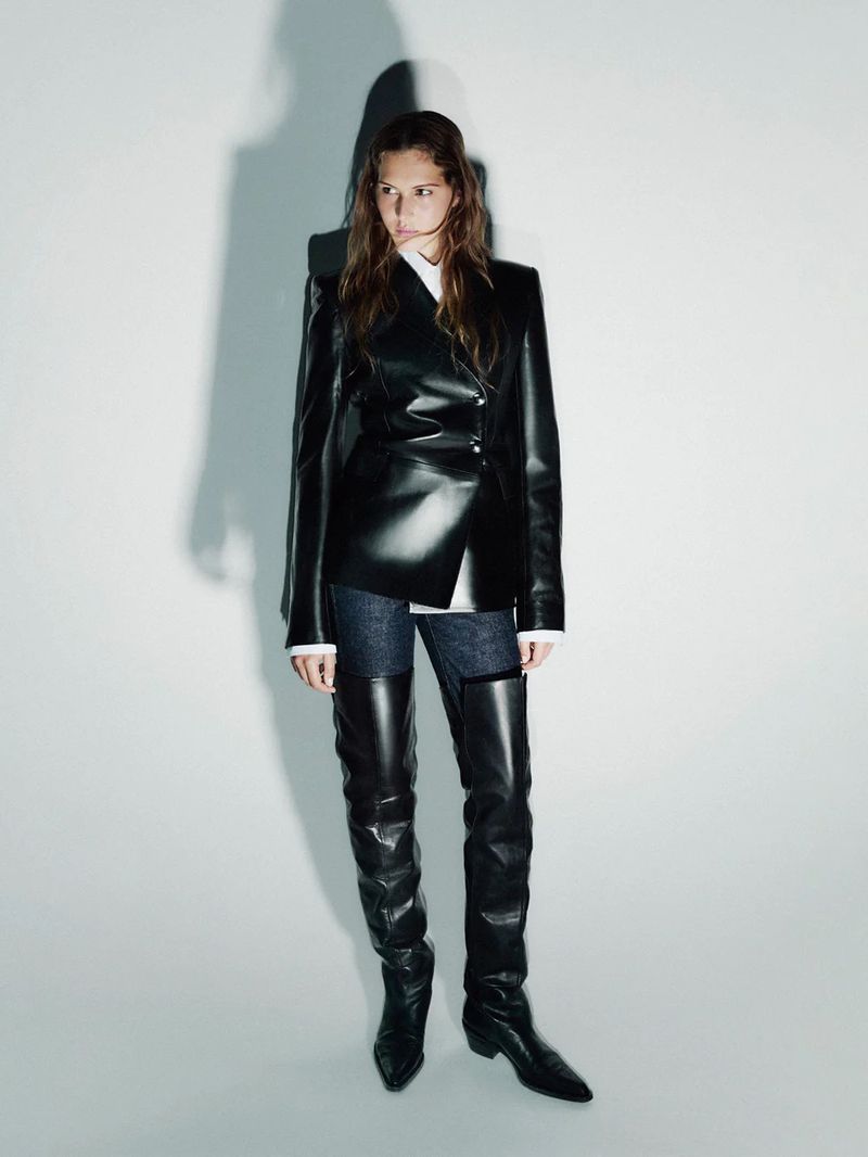 Fashionable woman in a black leather blazer and thigh-high boots poses in a studio with a neutral background and soft studio lighting.