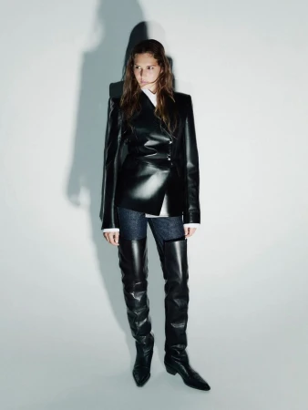 Fashionable woman in a black leather blazer and thigh-high boots poses in a studio with a neutral background and soft studio lighting.