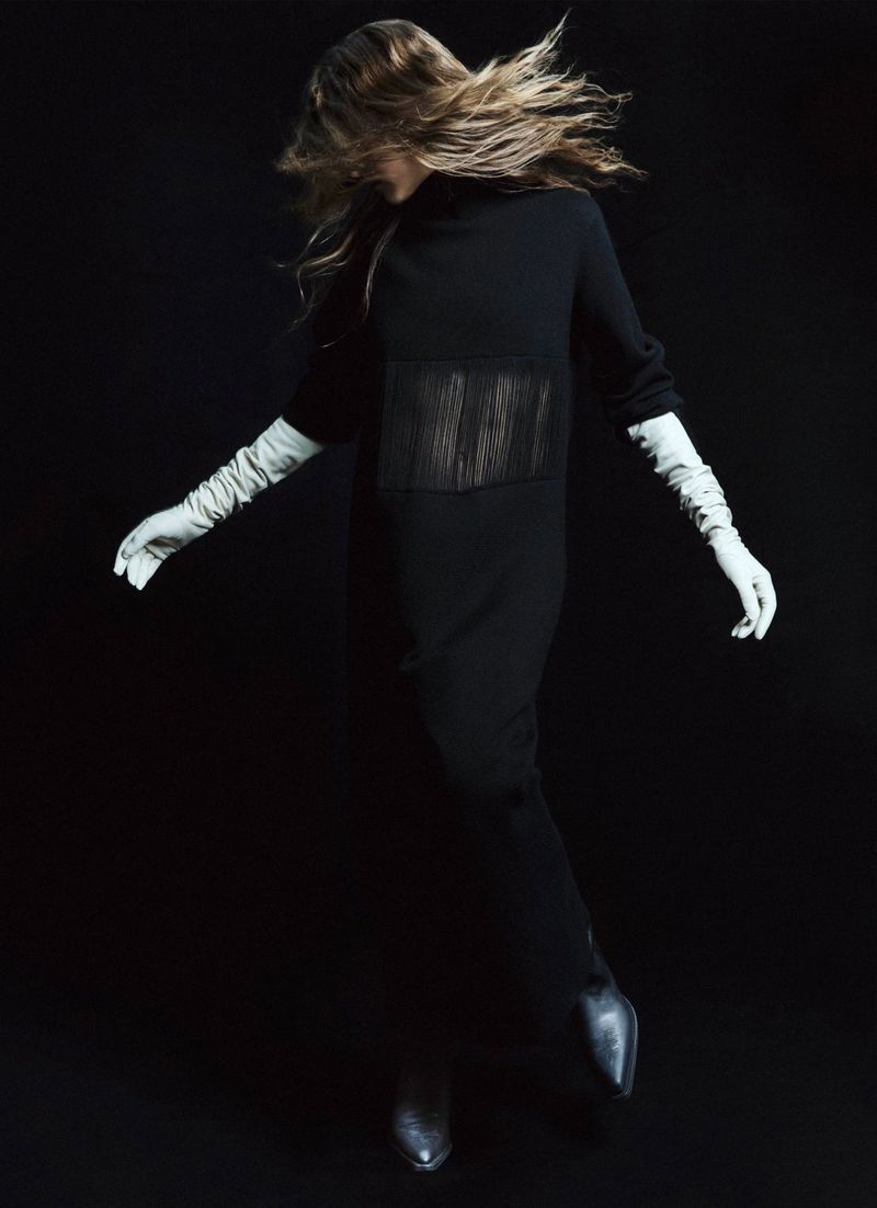 Fashion model wearing a black dress with sheer panel and white gloves poses in a dark studio environment with dynamic hair movement.