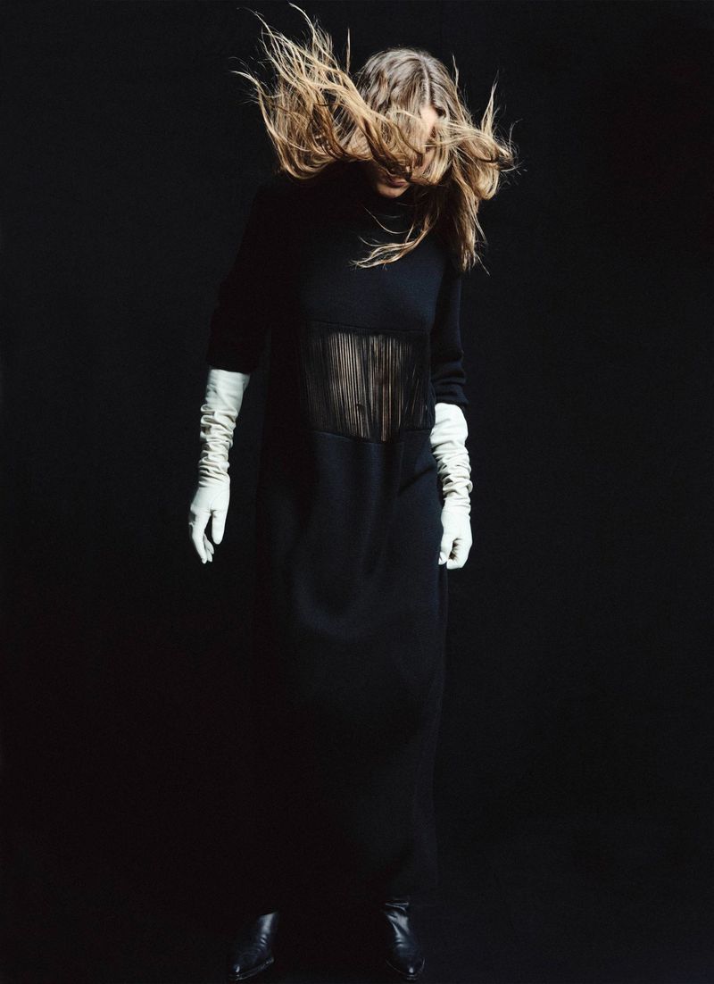 Fashion shot of an adult female wearing a black dress and long white gloves, posing in a dark photo studio with her hair in the air.