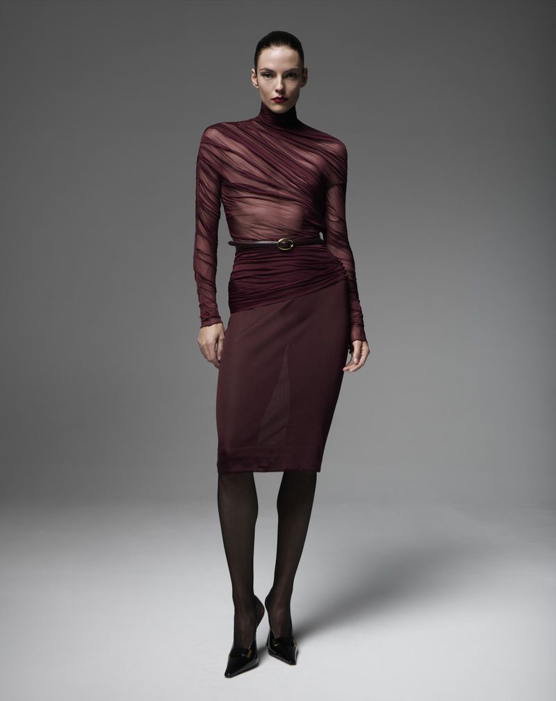Model Beauïse Genç wearing a sheer burgundy turtleneck top, pencil skirt, and black heels against a grey studio backdrop.