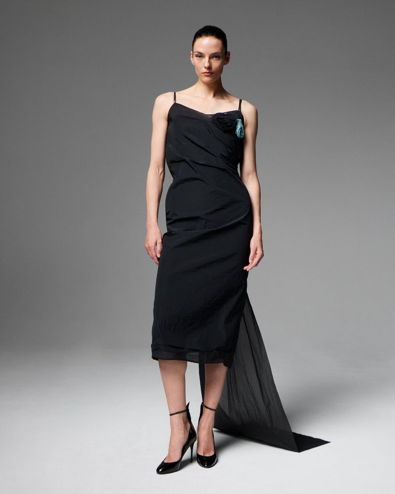 Elegant adult female fashion model poses in a black slip dress and high heels against a grey studio backdrop for a fashion editorial.