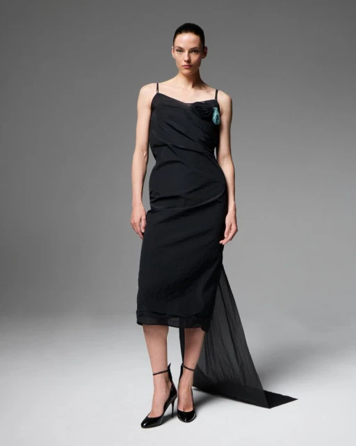 Elegant adult female fashion model poses in a black slip dress and high heels against a grey studio backdrop for a fashion editorial.