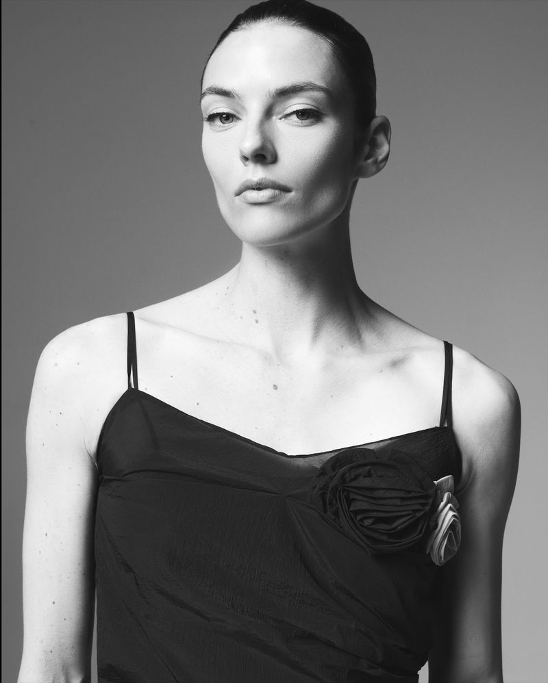 Elegant portrait of a fair-skinned adult female in a black dress with an embellished flower, set against a plain grey backdrop in the photo studio.