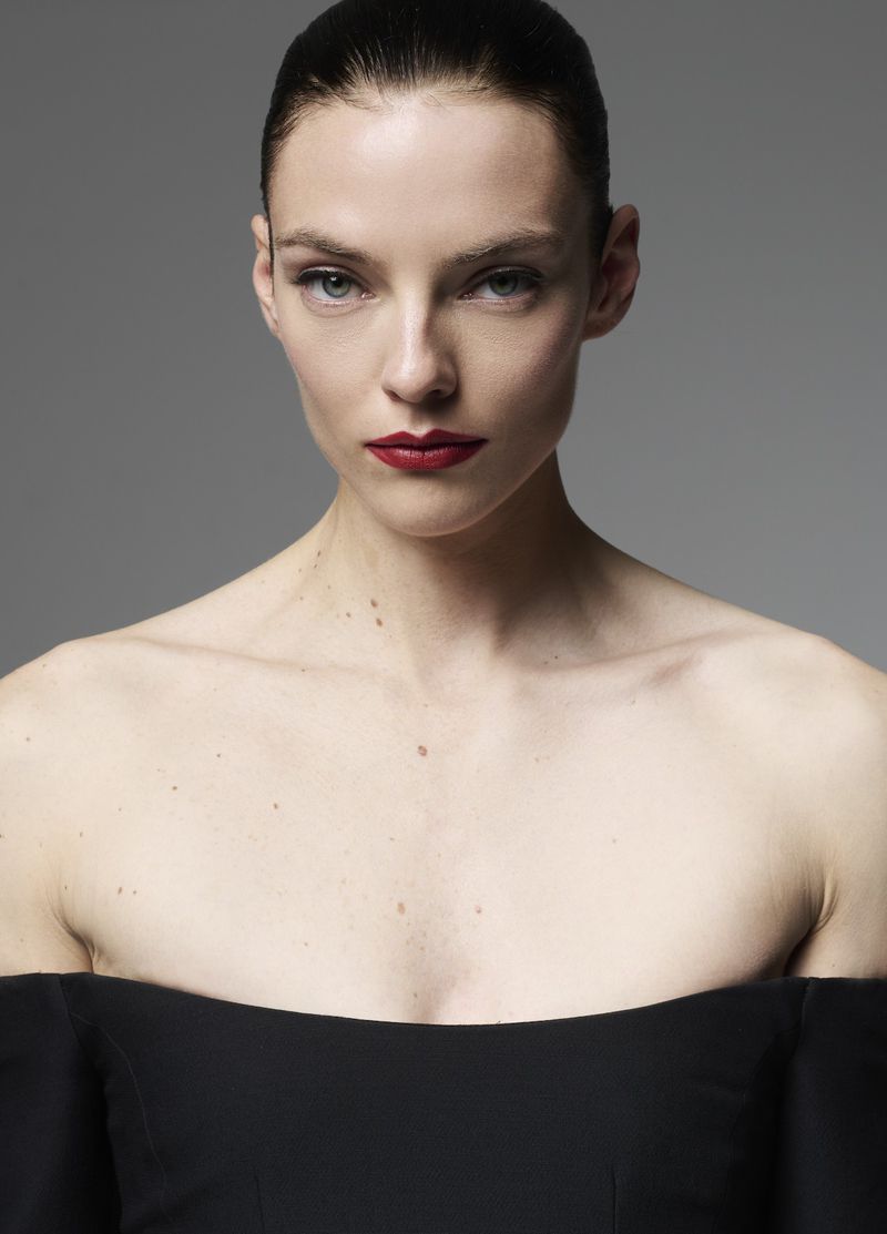 Portrait of a fair-skinned adult woman with red lipstick wearing a black off-the-shoulder top against a plain gray backdrop.