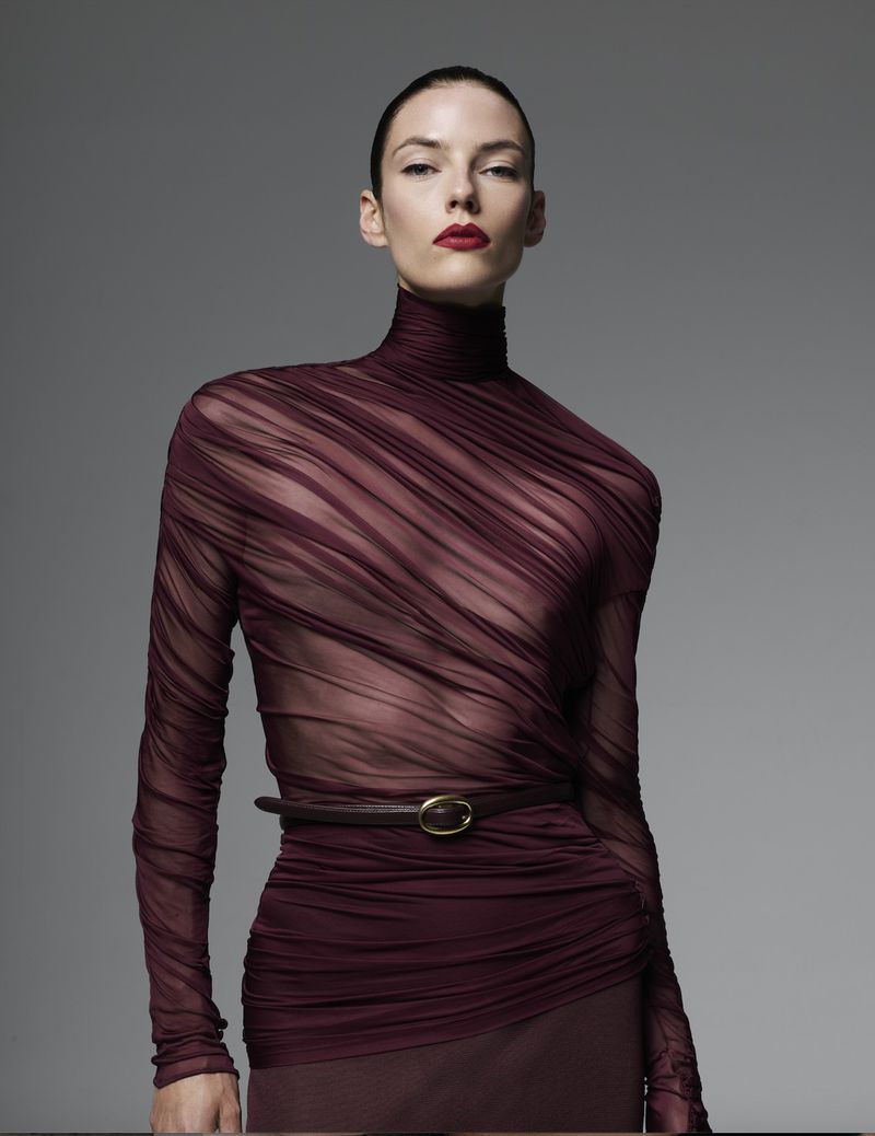 A striking portrait of a woman in a maroon, semi-sheer top with a brown belt and red lipstick, posing in front of a gray background.