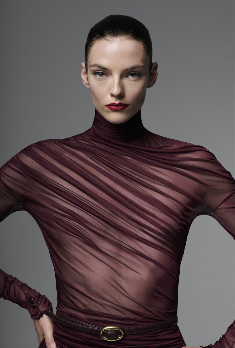 Fashion portrait of an adult woman wearing a sheer, maroon dress with a turtleneck and belt against a neutral gray backdrop.