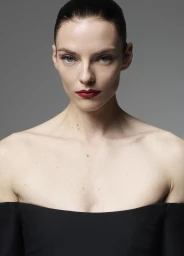 Model Beauïse Genç with slicked-back hair and bold red lipstick wearing a black off-the-shoulder top against a grey studio backdrop.