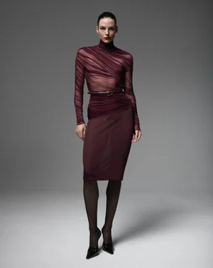 Model Beauïse Genç wearing a sheer burgundy turtleneck top, pencil skirt, and black heels against a grey studio backdrop.