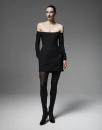 Model Beauïse Genç posing in an off-the-shoulder black mini dress with black tights and stiletto heels against a grey studio backdrop.