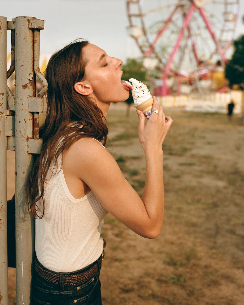 Woman enjoying a sprinkle-covered ice cream cone near a metal fence, with a Ferris wheel in the background during daytime.