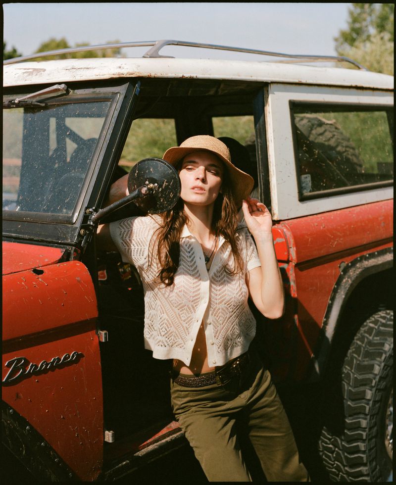 A woman in a hat poses next to a vintage red and white Bronco on a sunny day, leaning against the door, captured at eye-level.