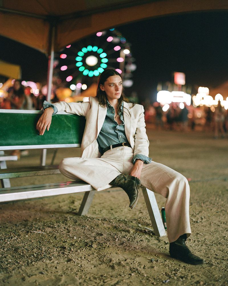 A woman in a blazer and wide-leg pants poses on a bench at night, with a Ferris wheel and carnival lights visible in the background.