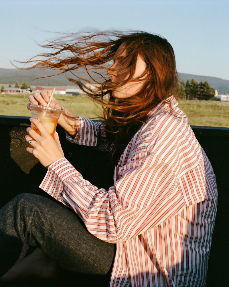 Woman in striped shirt drinks from plastic cup outdoors with windblown hair, sitting in back of vehicle with rural landscape in background.