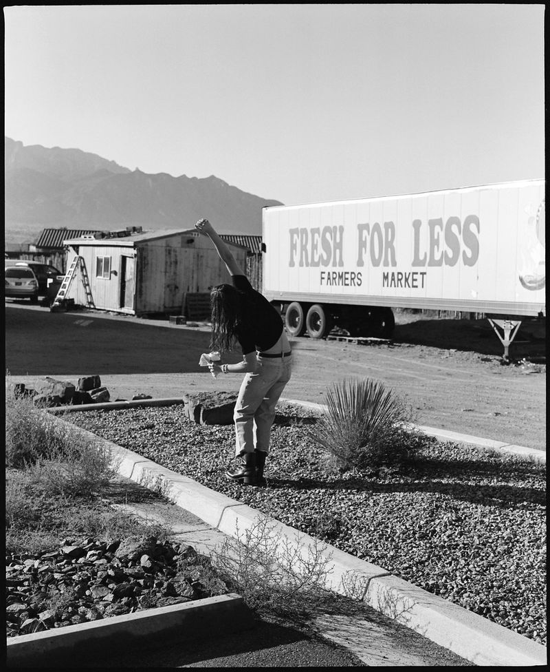 Black and white outdoor shot of a person with long dark hair in front of a Fresh For Less trailer with mountains in the background.