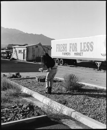 Black and white outdoor shot of a person with long dark hair in front of a Fresh For Less trailer with mountains in the background.