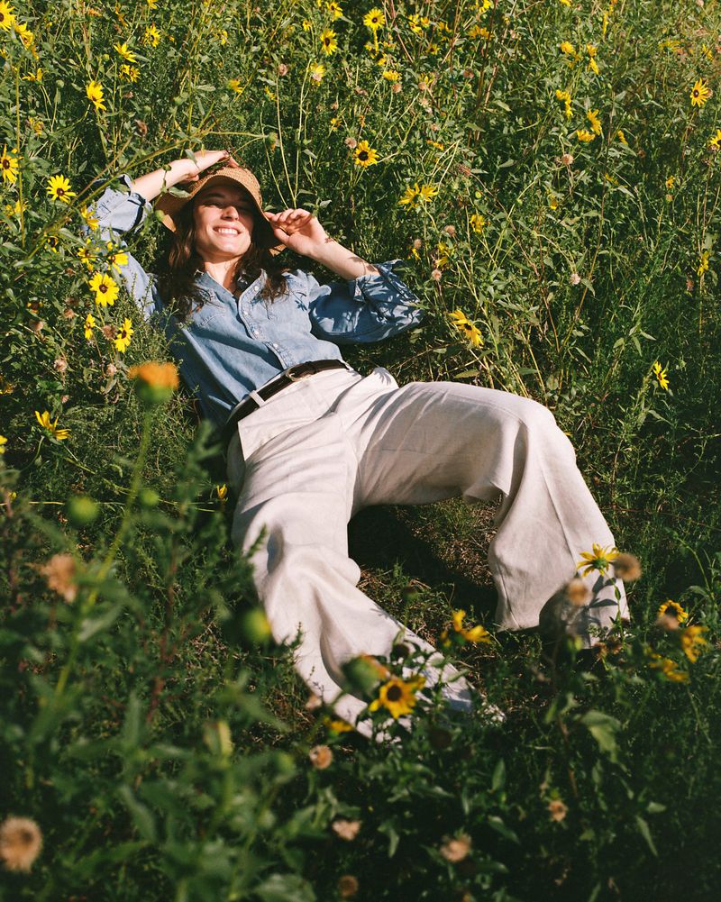A smiling woman in a hat relaxes in a field of wildflowers on a sunny day, wearing a light blue shirt and cream-colored pants.