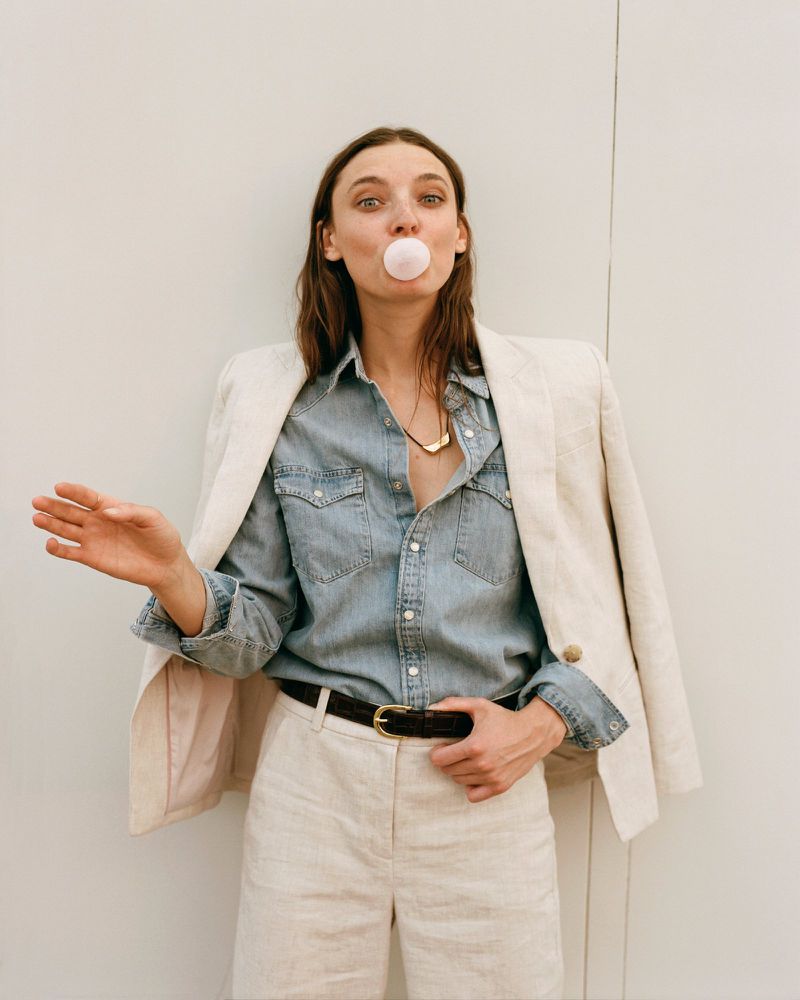 A young adult female poses outdoors in denim shirt and linen jacket and shorts, blowing a bubble with bubblegum and looking at the viewer.
