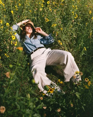 Ansley Gulielmi lying in a wildflower meadow wearing a denim shirt, white wide-leg trousers, and a sun hat.