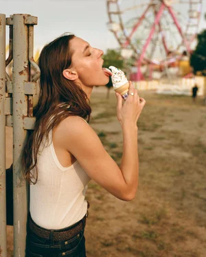 Ansley Gulielmi in a white tank top licking a soft-serve ice cream cone in front of a Ferris wheel.