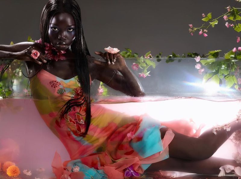 Stunning Black woman in a dress lies in water surrounded by flowers, against a dark backdrop for a fashion or beauty editorial.