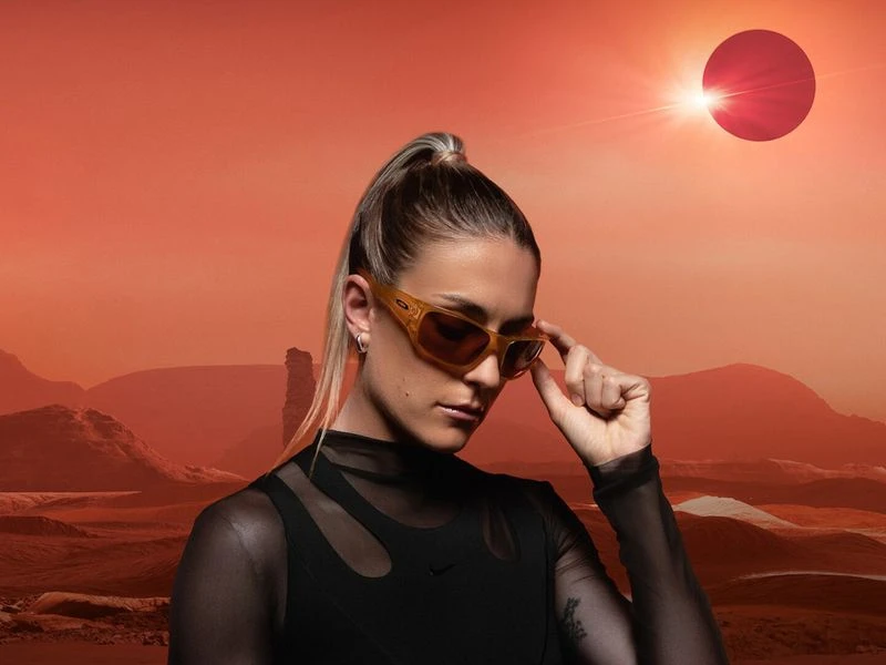 Woman in sunglasses poses in front of an orange desert landscape with a red planet or sun and mountains in the background.