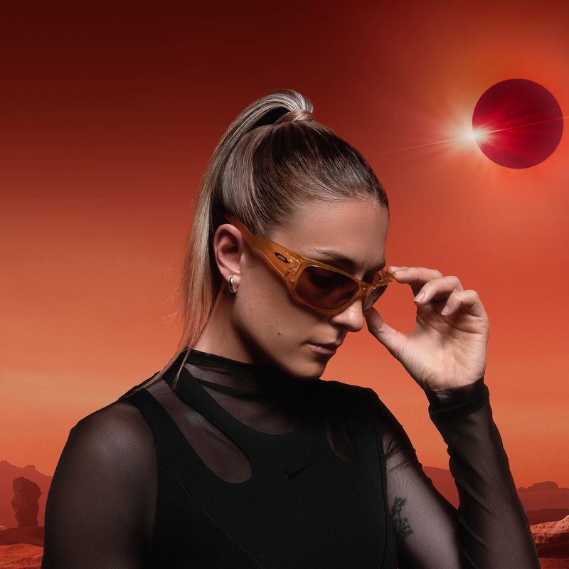 Portrait of a woman in sunglasses with a sleek ponytail, standing against a surreal orange and red landscape with a solar eclipse.