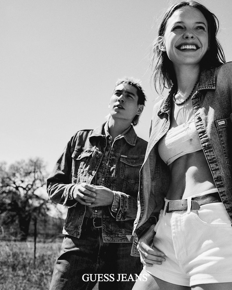A smiling woman and a man wear denim jackets in a black and white outdoor shot for a Guess Jeans fashion advertisement.