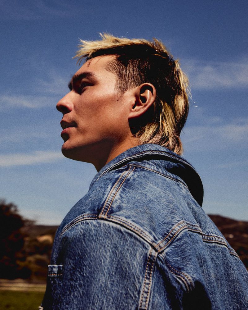 Portrait of a stylish adult man with a mullet wearing a denim jacket against a bright blue sky and grassy outdoor setting.