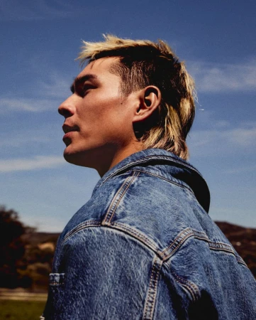 Portrait of a stylish adult man with a mullet wearing a denim jacket against a bright blue sky and grassy outdoor setting.