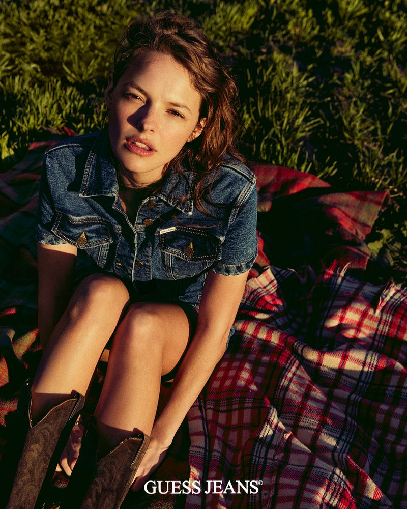 Attractive woman poses in Guess Jeans denim jacket, sitting on a plaid blanket outdoors in green grass. The fashion shot features fall colors.