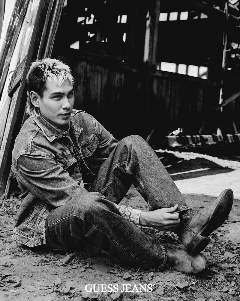 Monochrome portrait of a person in a denim jacket and jeans sitting on the ground in front of a dilapidated wooden structure.