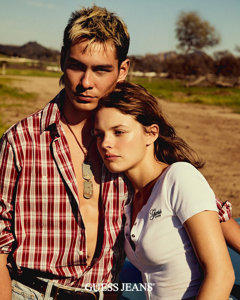 Fashion image of a young adult couple outdoors, wearing casual clothing. The man wears a plaid shirt, and the woman a white top.