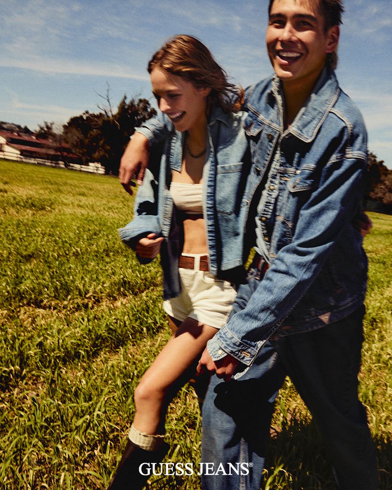 Smiling young couple in denim jackets pose together in a grassy field under a sunny sky in a fashion advertisement for Guess Jeans.