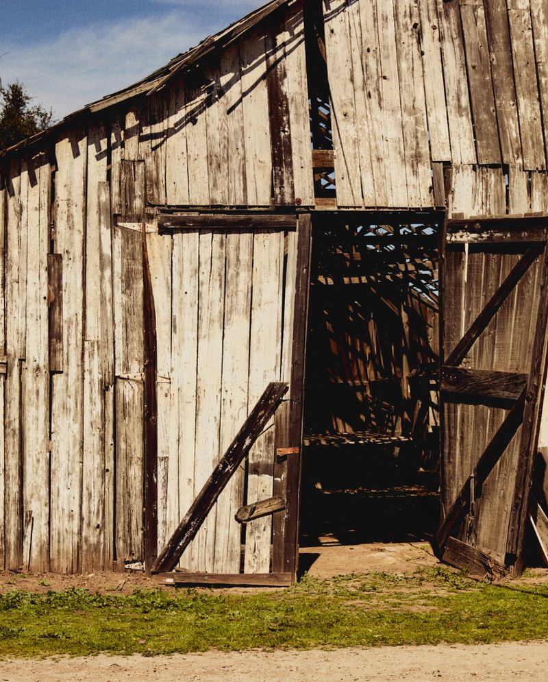 Exterior view of an old, weathered barn with peeling white paint and an open door, set against a backdrop of green grass and blue sky.