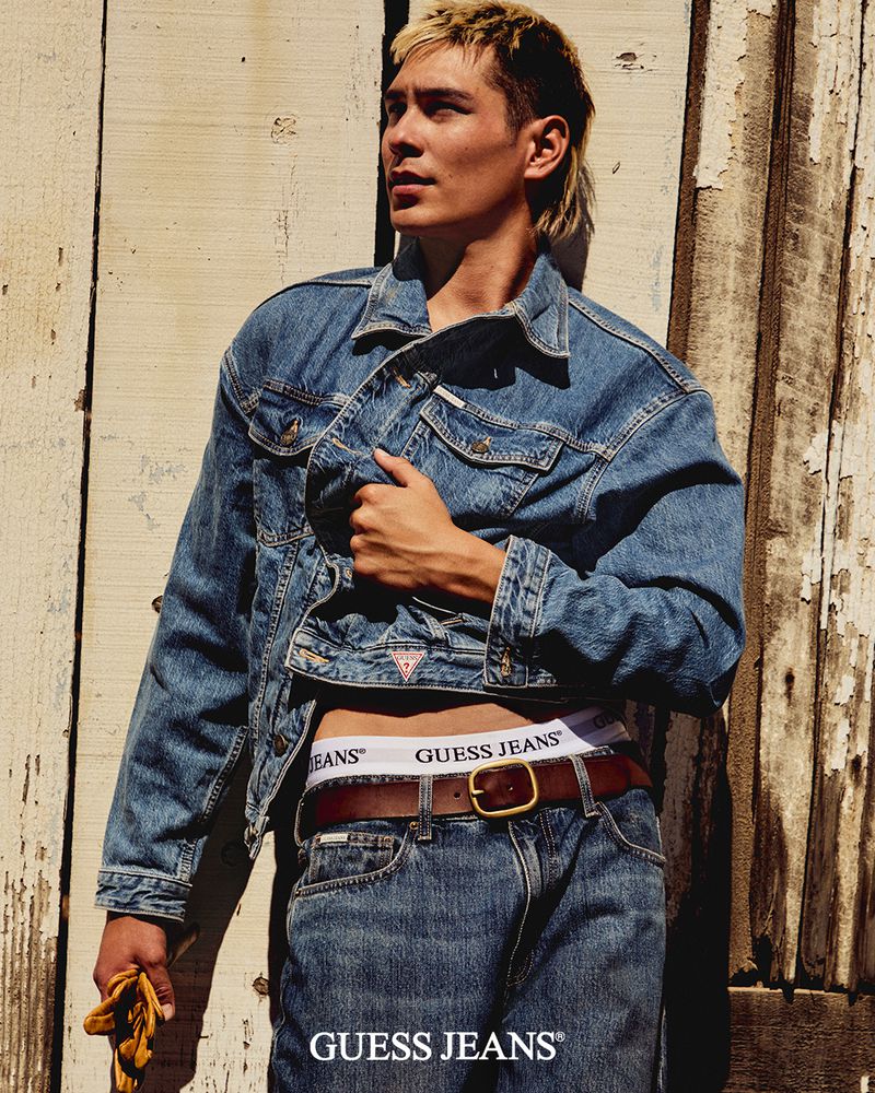 Young adult male in denim jacket and jeans poses against a weathered wood background. He has a blond mullet hairstyle and a brown belt.