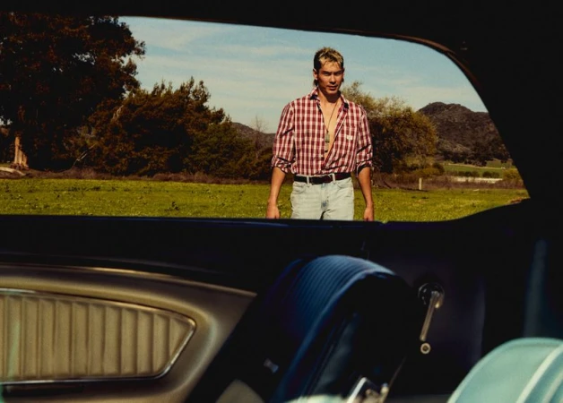 Portrait of a young man in a red plaid shirt and blue jeans standing in a field, shot from inside a car with leather interior.